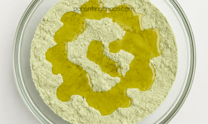 Green moon sand mixture in a bowl with oil poured on top before mixing.