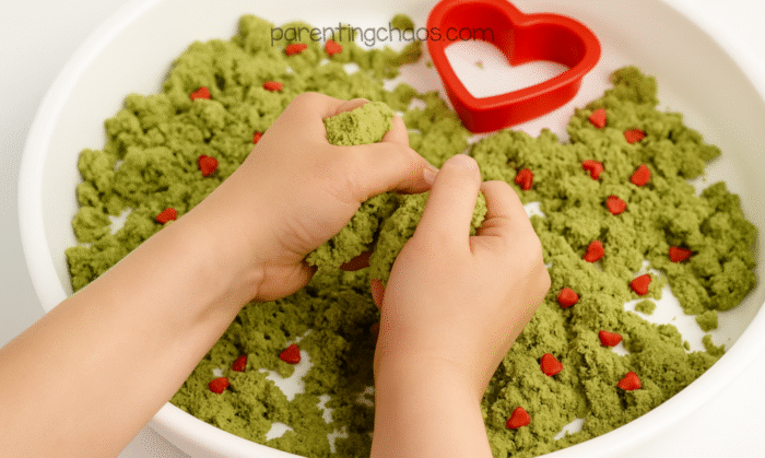 A child’s hands squeezing and playing with green Grinch moon sand in a sensory tray with red heart sprinkles.