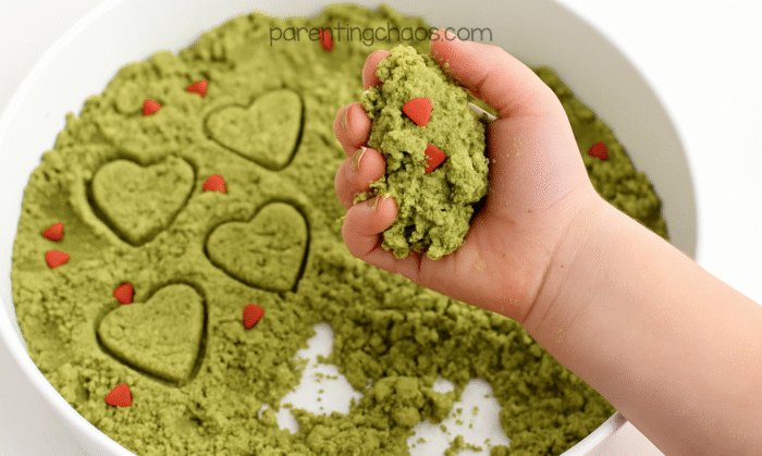 A child holding a handful of green Grinch moon sand with red heart sprinkles, with heart shapes stamped in the tray.