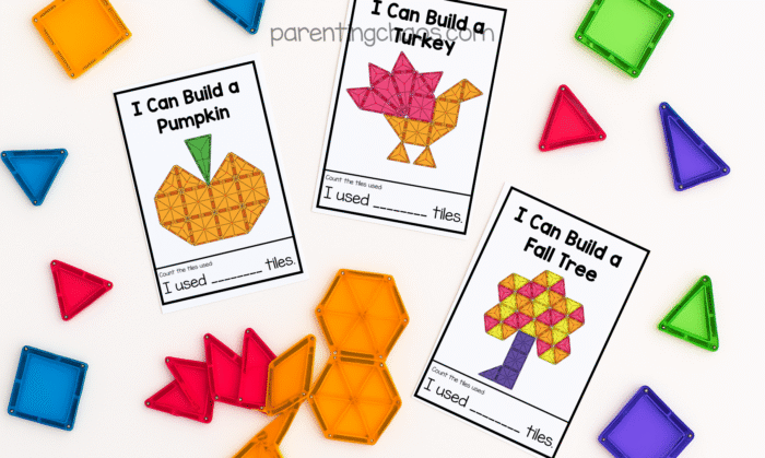 Overhead view of three Thanksgiving magnetic tile task cards on a white background. The cards read “I Can Build a Pumpkin,” “I Can Build a Turkey,” and “I Can Build a Fall Tree,” each with a colorful design made from translucent magnetic tiles. Loose tiles in red, orange, blue, green, and yellow surround the cards.